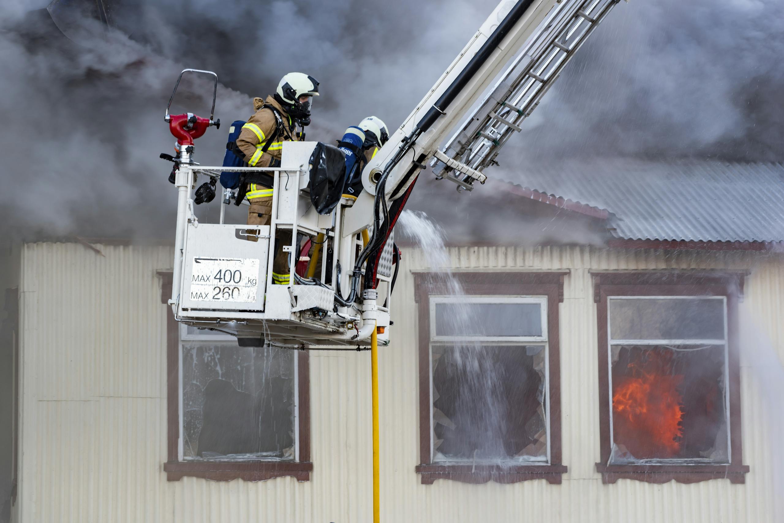 Firefighters on a ladder extinguishing a house fire with smoke and flames visible.